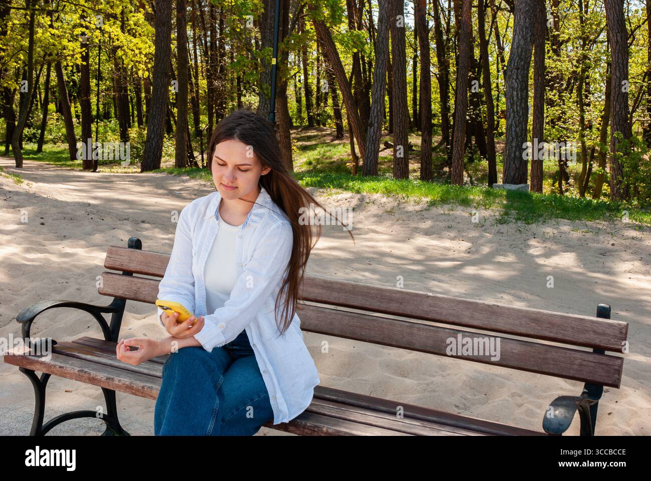 Jeune femme avec de longs cheveux bruns assis sur un banc en bois dans un parc de sable ensoleillé, à l'aide d'un smartphone et profitant d'un moment de détente dans la nature, symboli Banque D'Images