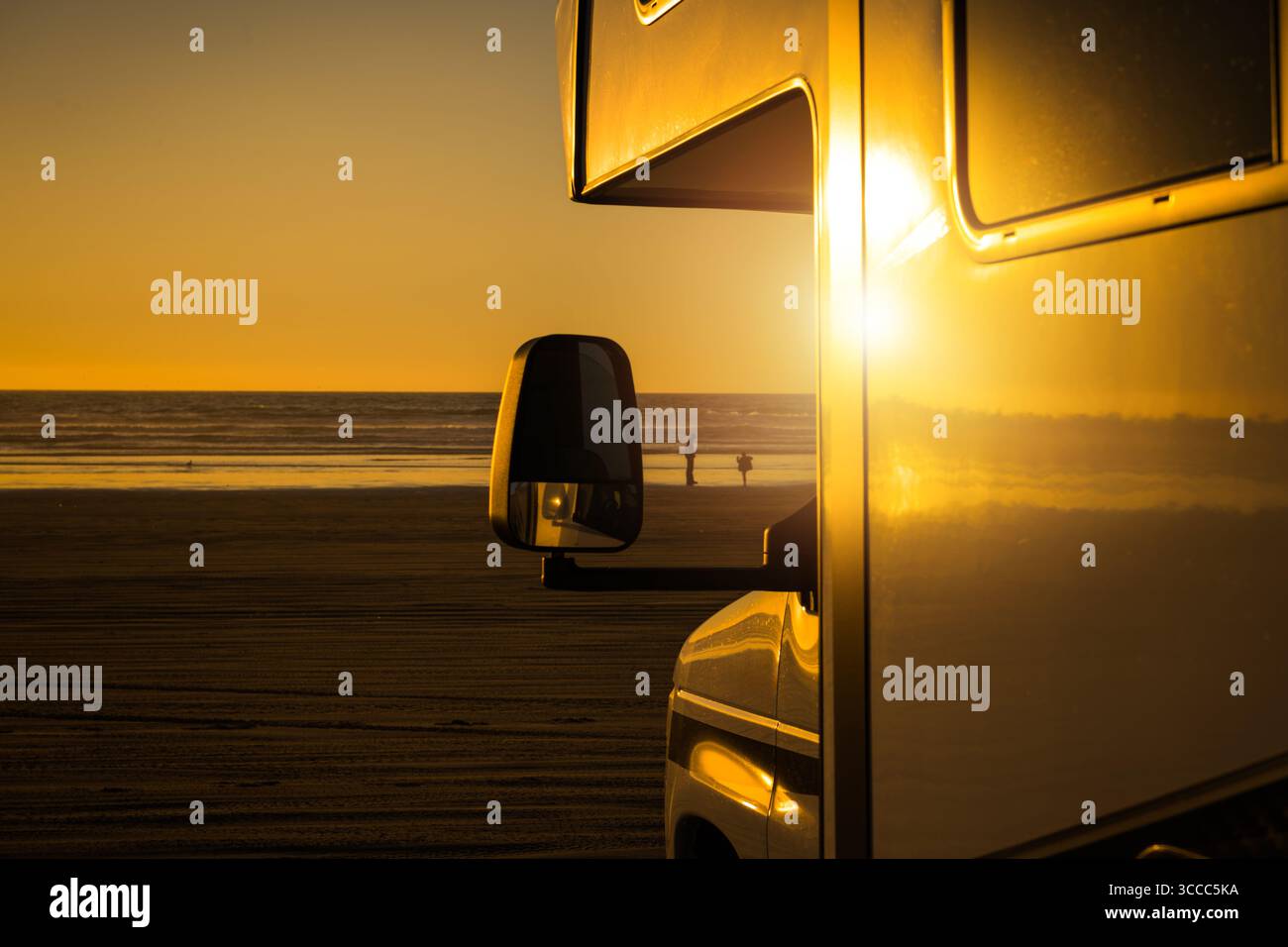 Un camping-car se tient près du rivage pendant le coucher du soleil, avec la lumière dorée qui se reflète sur sa surface tandis que les gens se promènent le long de la plage dans le backgrou Banque D'Images