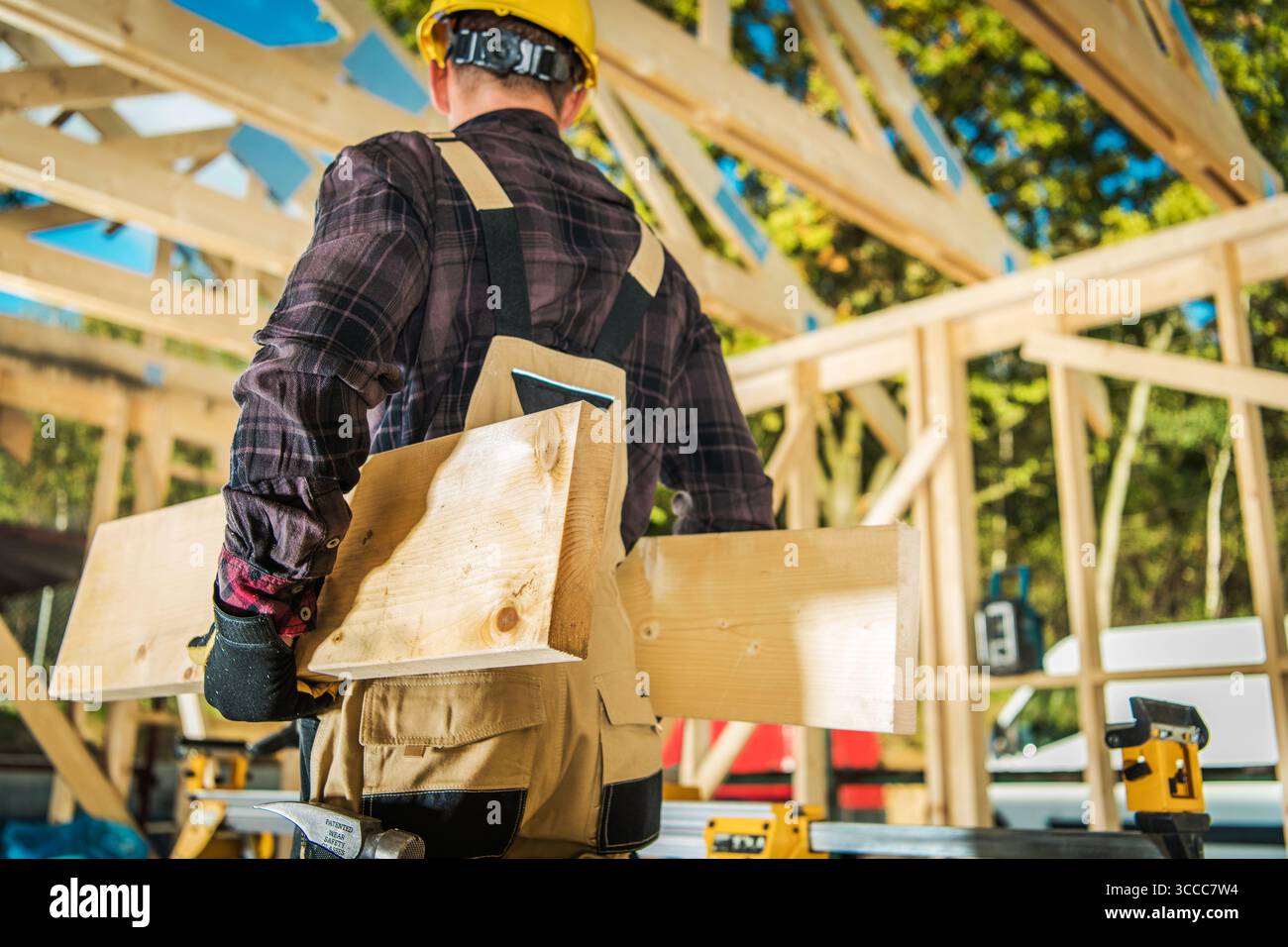 Un ouvrier de la construction portant un casque et des gants transporte des planches de bois sur un chantier ensoleillé, mettant en valeur la phase active des travaux de construction. Banque D'Images