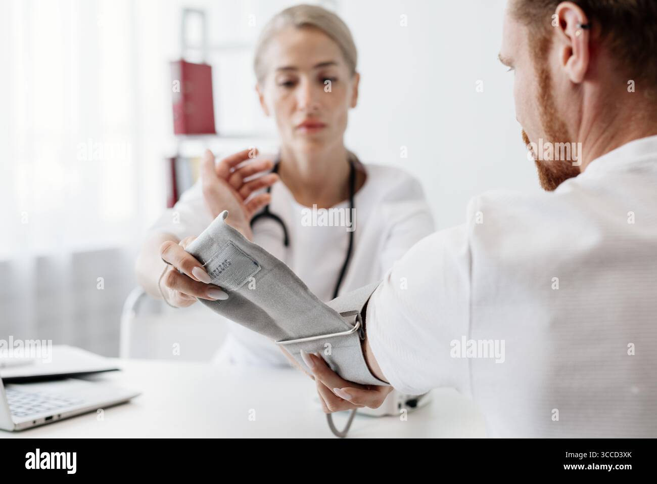 Médecin féminin mesure la pression artérielle d'un patient masculin dans une clinique moderne, avec du matériel médical et un ordinateur portable sur le bureau, mettant l'accent sur les profes de soins de santé Banque D'Images