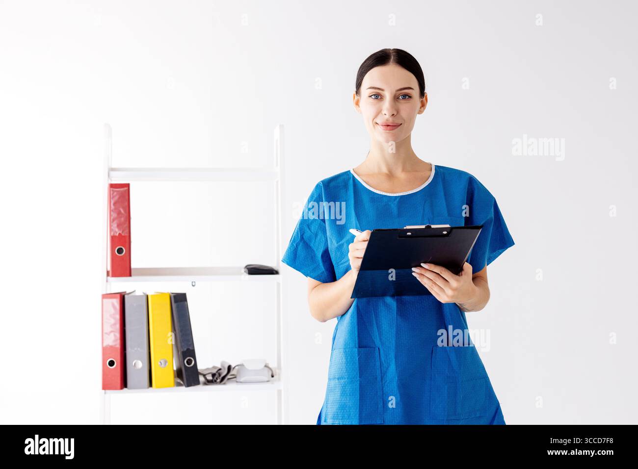 Professionnel de santé féminin dans les gommages bleus, tenant une planche à pince, se tient dans une clinique moderne avec des étagères organisées, démontrant l'engagement envers le patient Banque D'Images