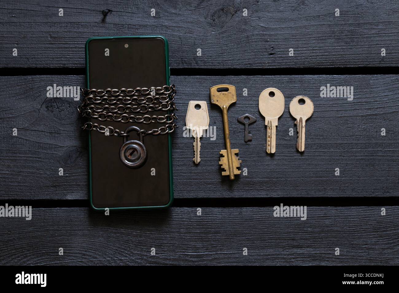Smartphone dans un étui vert enveloppé d'une chaîne métallique avec un cadenas, cinq clés différentes sur une surface en bois noir Banque D'Images