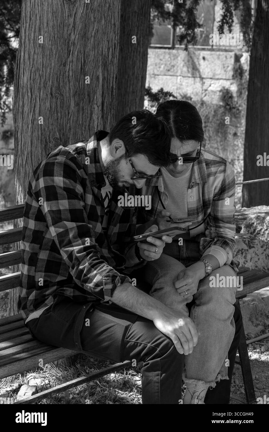 Un couple est assis sur un banc à l'Acropole, Athènes, regardant leur smartphone par une chaude journée d'été, la ville ensoleillée et les ruines antiques autour d'eux. Banque D'Images