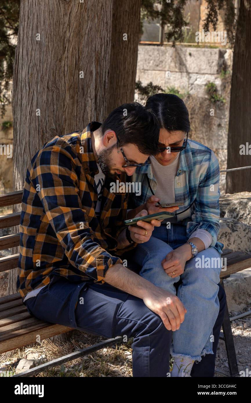 Un couple est assis sur un banc à l'Acropole, Athènes, regardant leur smartphone par une chaude journée d'été, la ville ensoleillée et les ruines antiques autour d'eux. Banque D'Images