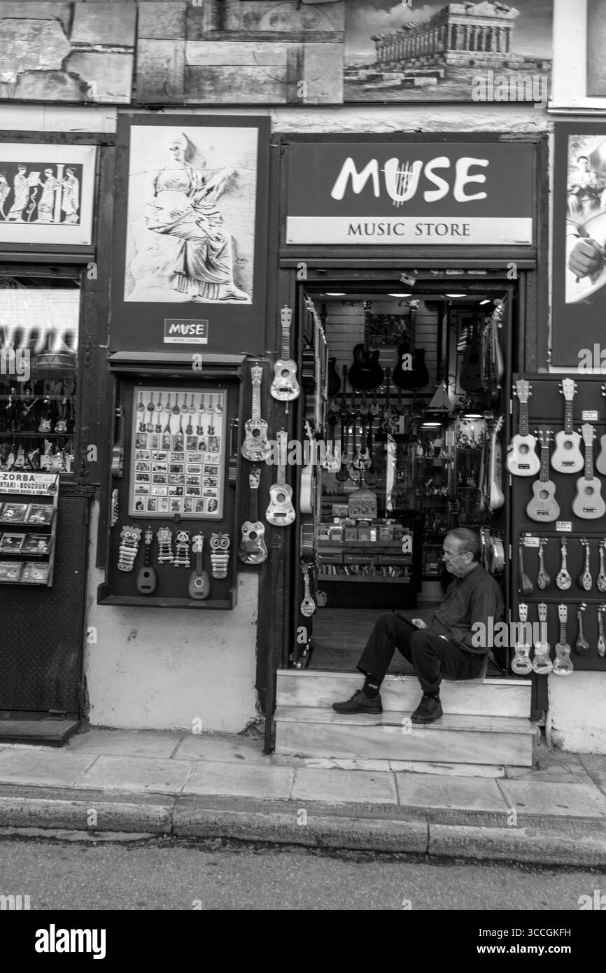 Un homme est assis dans la porte d'un magasin de musique encombré à Athènes, en Grèce, entouré d'instruments par une journée d'été ensoleillée, la vie urbaine autour de lui. Banque D'Images