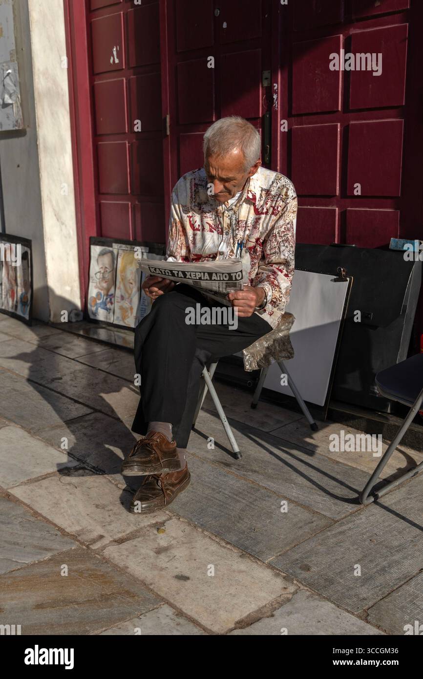 Un homme est assis sur un banc usé à Athènes, en Grèce, lisant un journal sous le soleil d'été, entouré par les rues de la ville, le béton et le squaller Banque D'Images