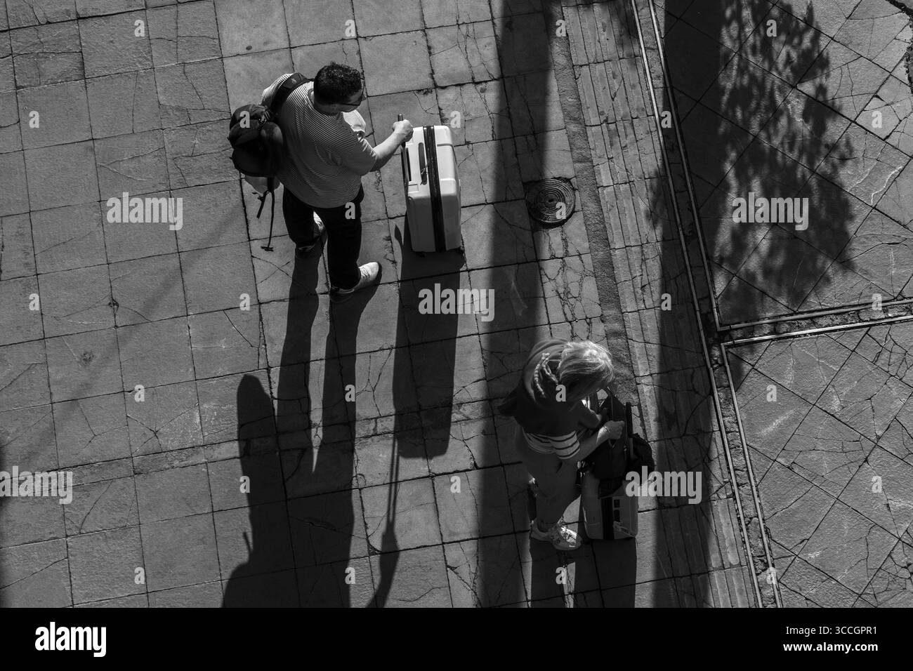 Deux voyageurs avec des bagages enregistrent leurs bagages dans une rue d'Athènes, vue d'en haut par une fenêtre d'hôtel par une chaude journée d'été sous la lumière du soleil. Banque D'Images