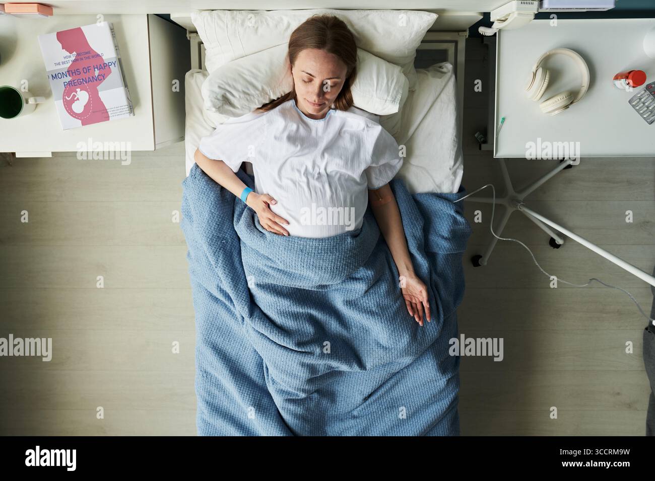 Femme jeune adulte enceinte caucasienne couchée dans un lit d'hôpital reposant la main sur le ventre, équipement médical et livre de maternité visible sur la table de chevet, vue de dessus Banque D'Images