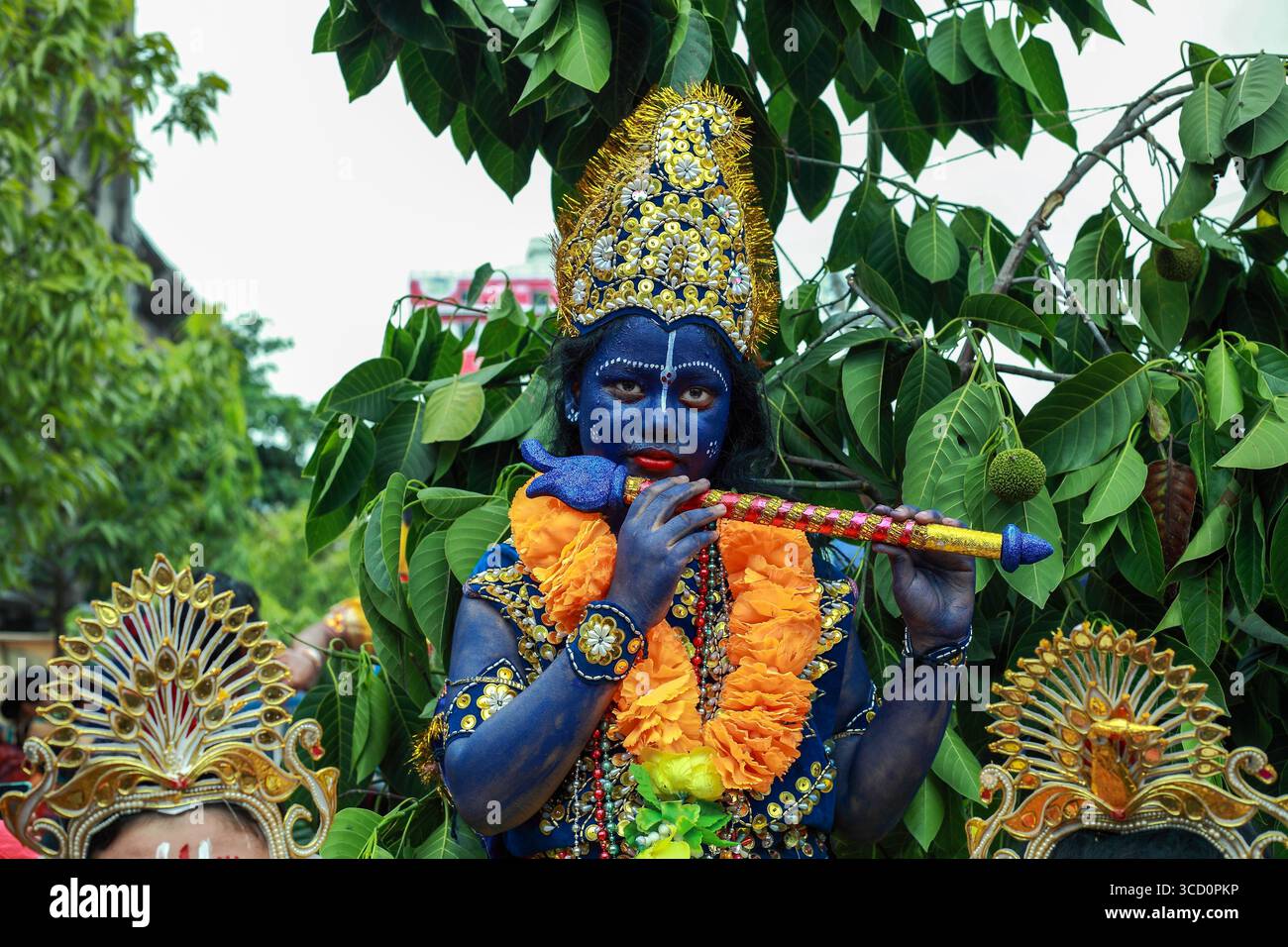 Narayanganj, Bangladesh - 23 août 2019 : vue d'une personne ornée d'un maquillage bleu vif et d'une couronne dorée, serenading avec une flûte au milieu d'un éclat luxuriant Banque D'Images