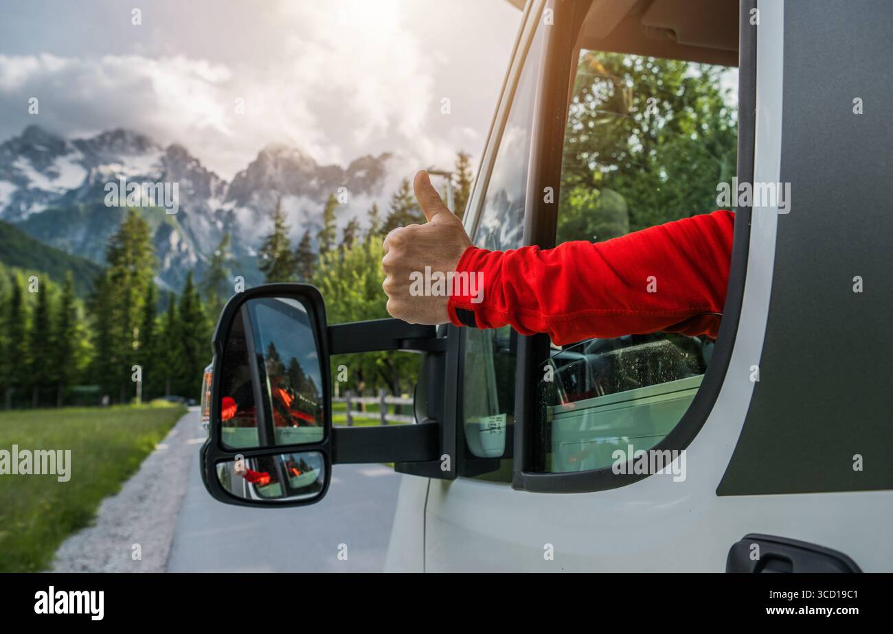 Un voyageur dans une chemise rouge à manches longues donne un pouce vers le haut de la fenêtre d'une camionnette tout en conduisant à travers un paysage de montagne pittoresque au coucher du soleil. Banque D'Images