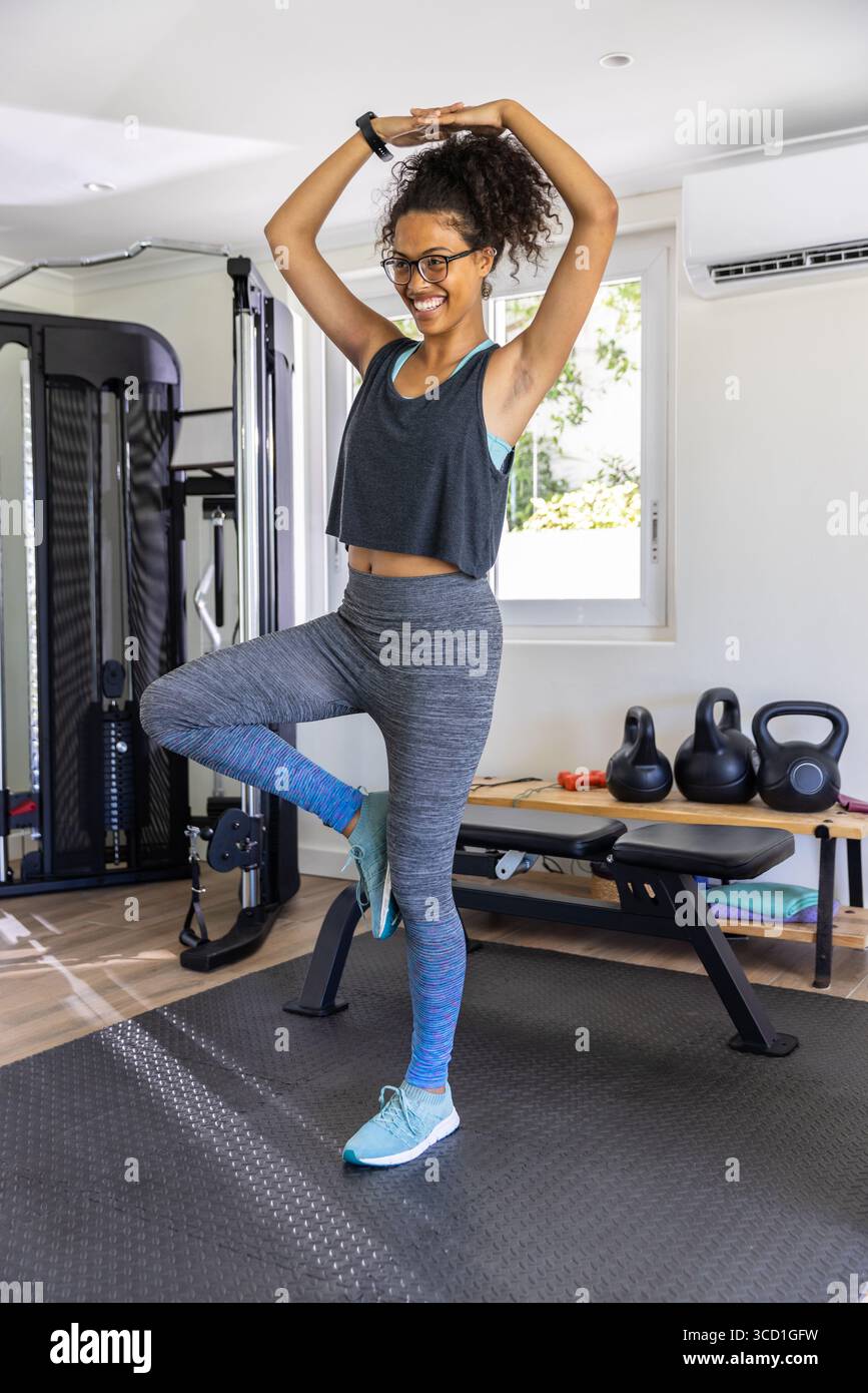 Femme afro-américaine pratiquant l'arbre de yoga pose dans le gymnase sur tapis près de la machine de câble et kettlebells Banque D'Images