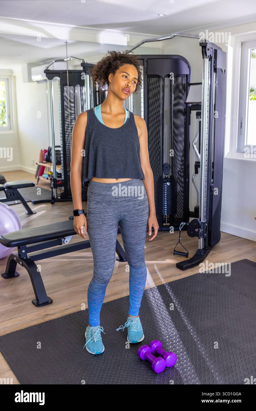 Femme afro-américaine exerçant avec des haltères violets sur tapis noir dans la salle de gym à domicile avec mur miroir Banque D'Images