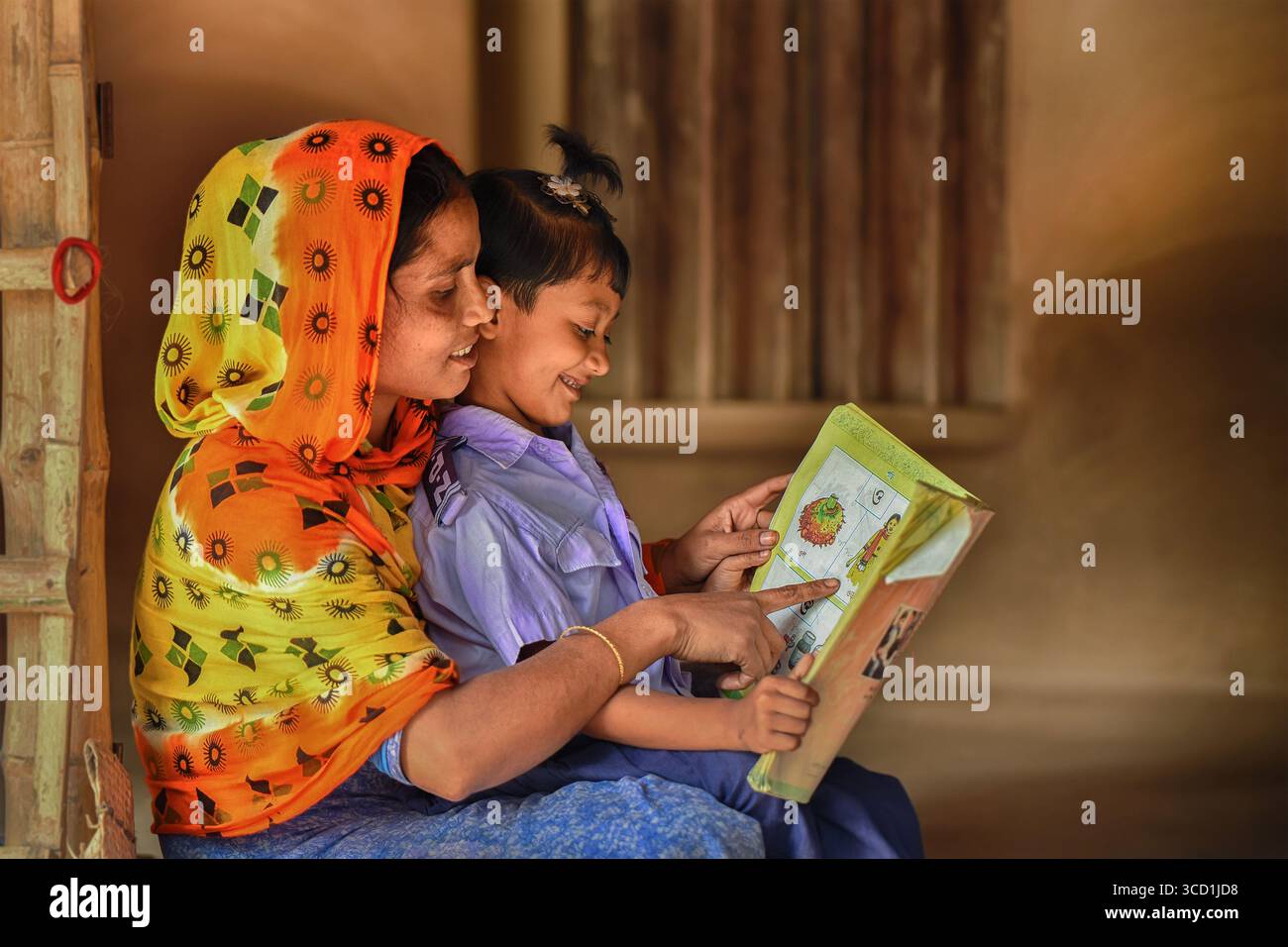 Bogura, Bangladesh - 15 décembre 2019 : vue d'une mère et d'un enfant, baignés de lumière douce, partageant un livre coloré sur fond de maison simple. Banque D'Images