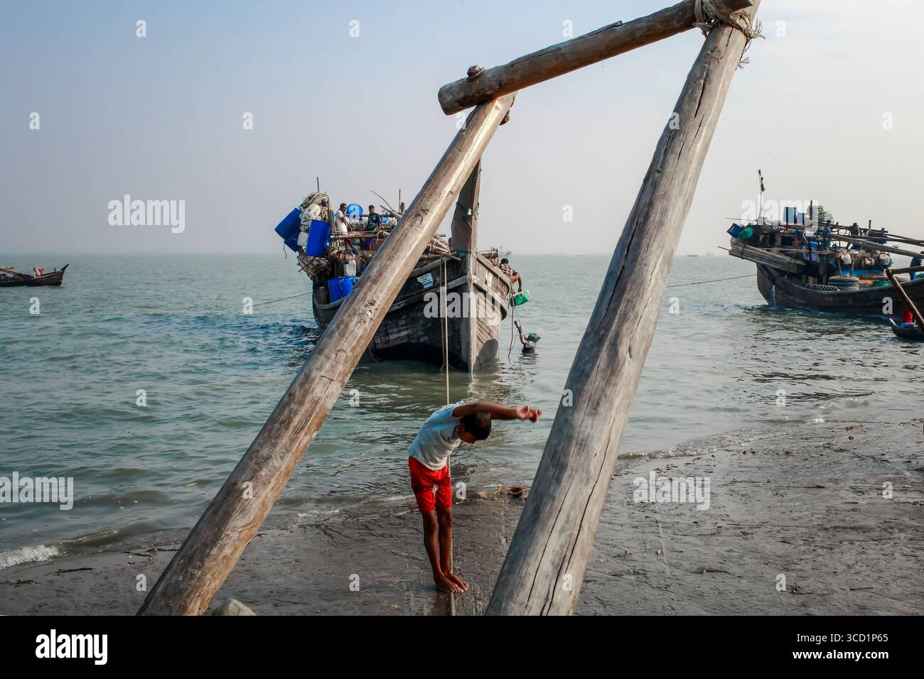 Cox's Bazar, Bangladesh - 07 mars 2022 : vue d'un garçon debout devant des structures en bois altérées, avec des bateaux de pêche reposant sur la rive scintillante sous un ciel pâle. Banque D'Images
