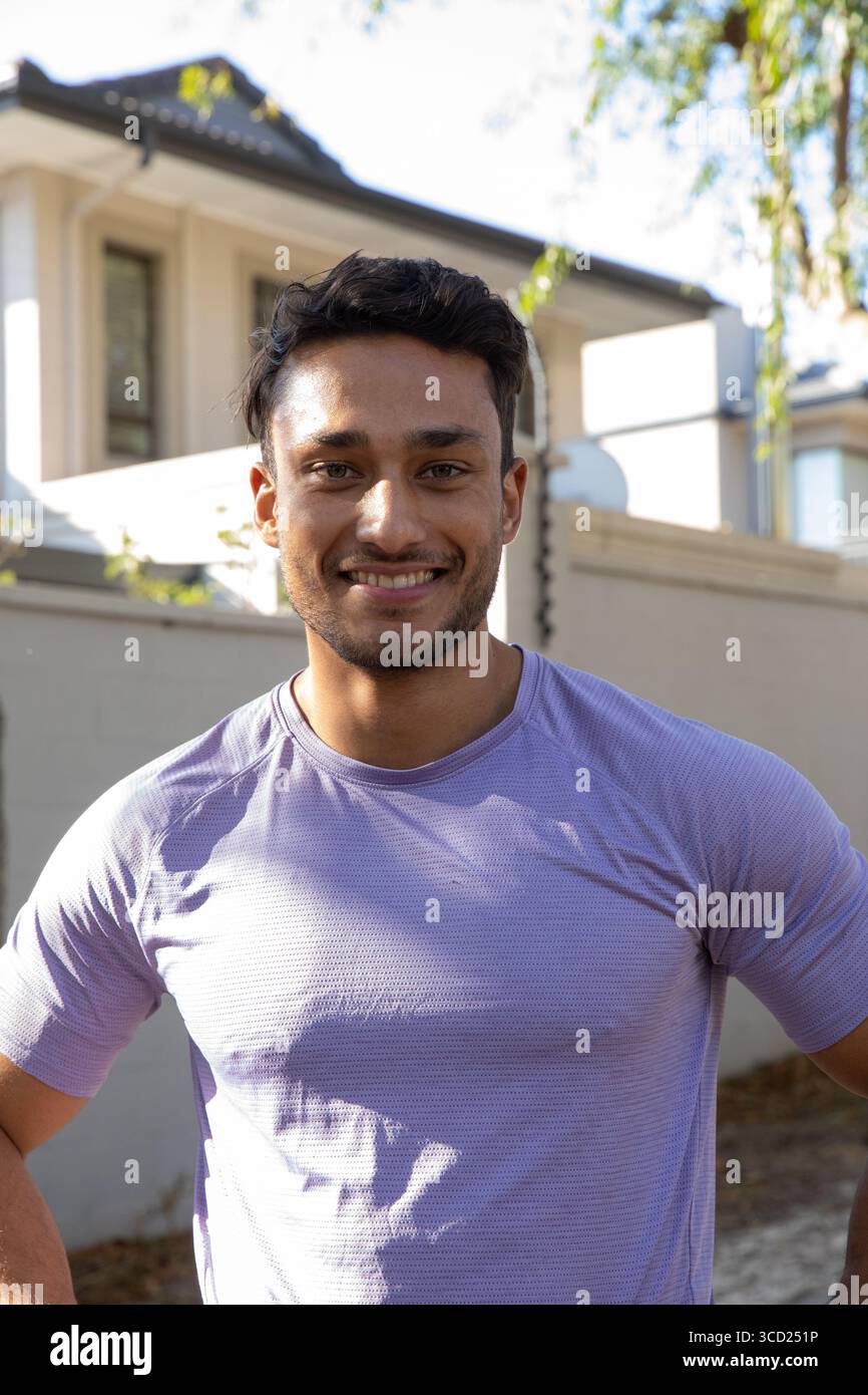 Homme indien debout souriant devant l'extérieur de la maison de banlieue avec bas mur de briques sous la lumière du soleil Banque D'Images