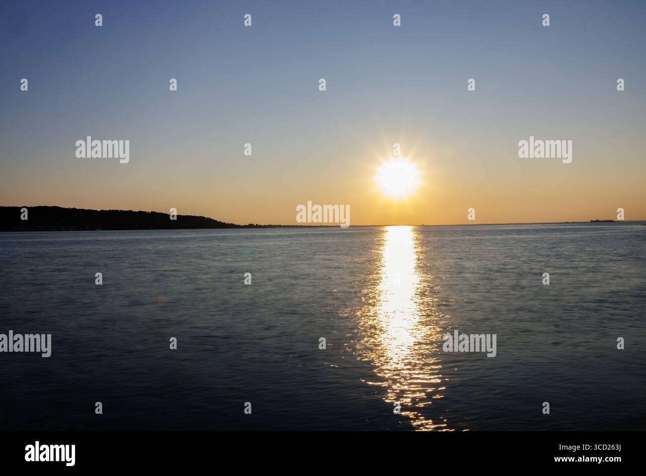 Soleil brillant sur les eaux calmes de la mer avec un ciel bleu clair et reflet doré menant à la côte lointaine dans la scène côtière paisible. Banque D'Images