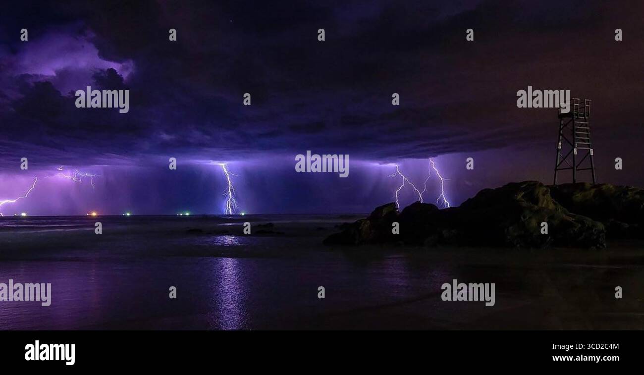 La vue des coups de foudre illumine le ciel sombre au-dessus de l'océan, se reflétant dans le sable humide près d'une tour de sauveteurs, Redhead Beach, Newcastle, Australie. Banque D'Images