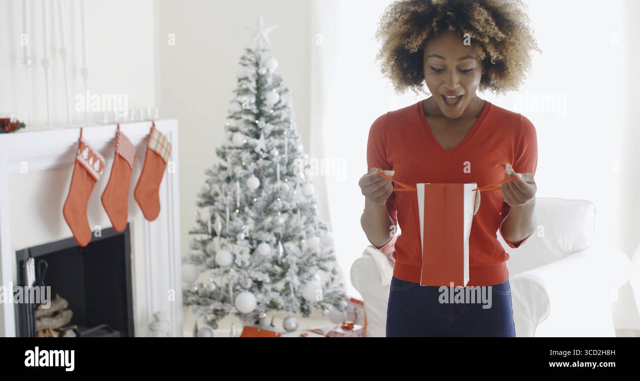 Heureuse jeune femme africaine attrayante ouvrant des cadeaux de Noël devant l'arbre regardant dans un sac cadeau dans la surprise Banque D'Images