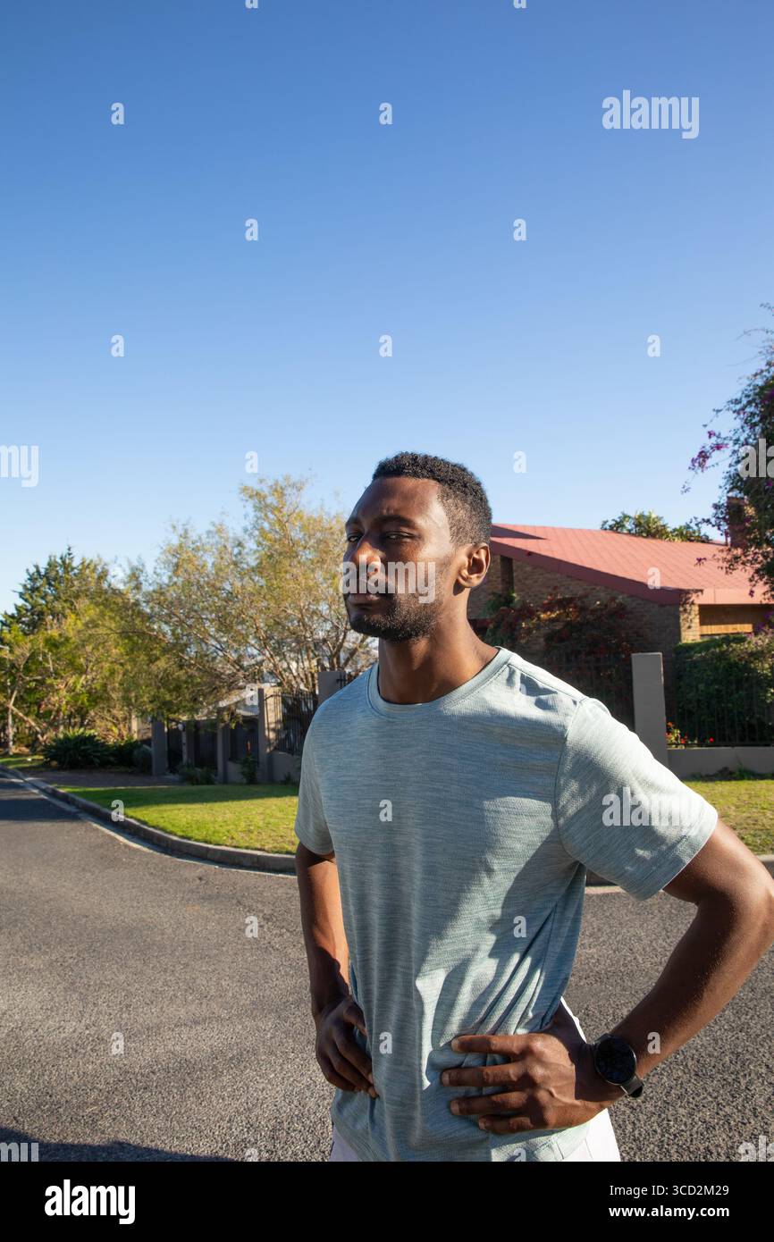 Homme afro-américain faisant une pause à mi-course sur la rue de banlieue sous le ciel clair tout en vérifiant la montre de sport Banque D'Images