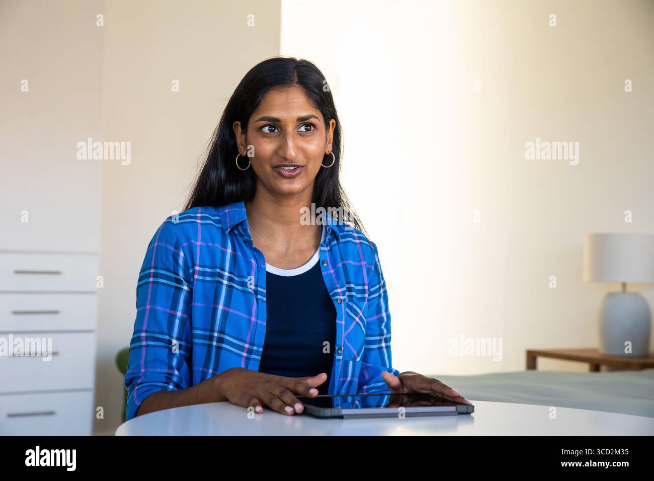 Femme indienne utilisant la tablette tout en étant assis à la table blanche près de la lampe en céramique grise dans la pièce lumineuse Banque D'Images