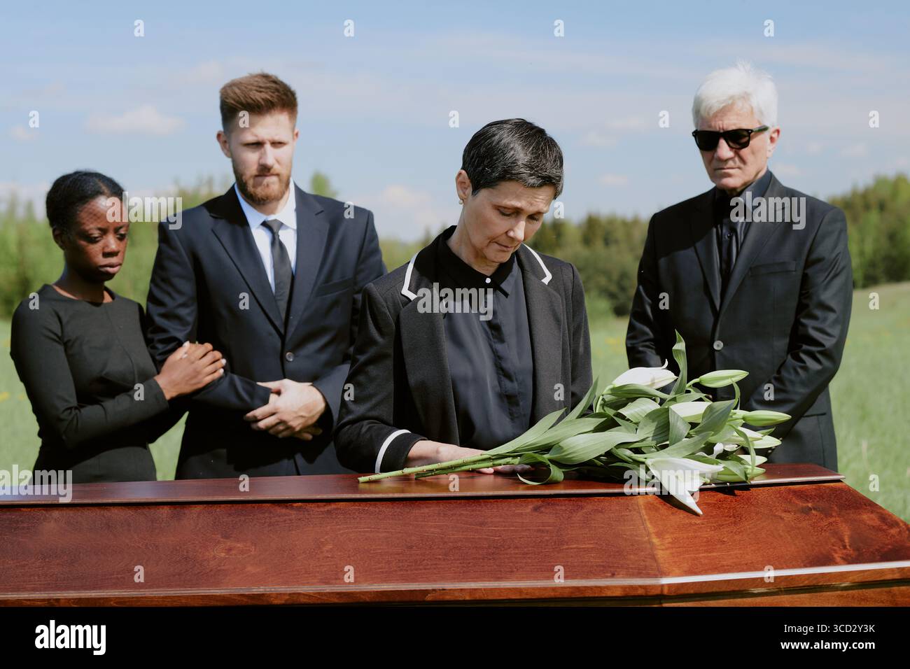 Femme caucasienne d'âge moyen tenant des lis debout près d'un cercueil en bois à l'extérieur, femme noire et homme caucasien debout ensemble, homme caucasien senior portant des lunettes de soleil debout à proximité Banque D'Images
