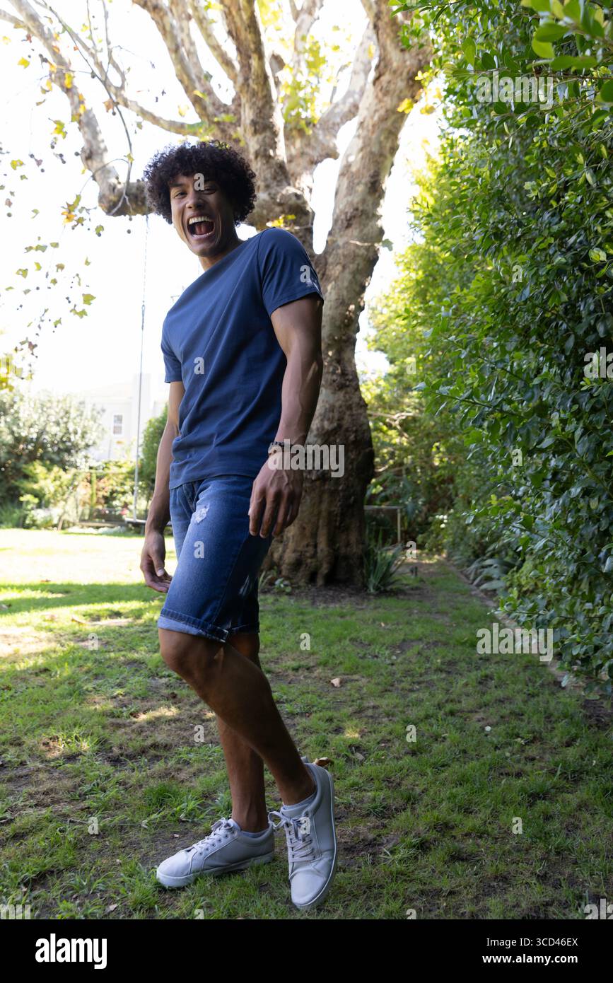 Homme riant en se penchant sur le tronc d'arbre dans la cour près de la haie verte, bracelet perlé et corde Banque D'Images