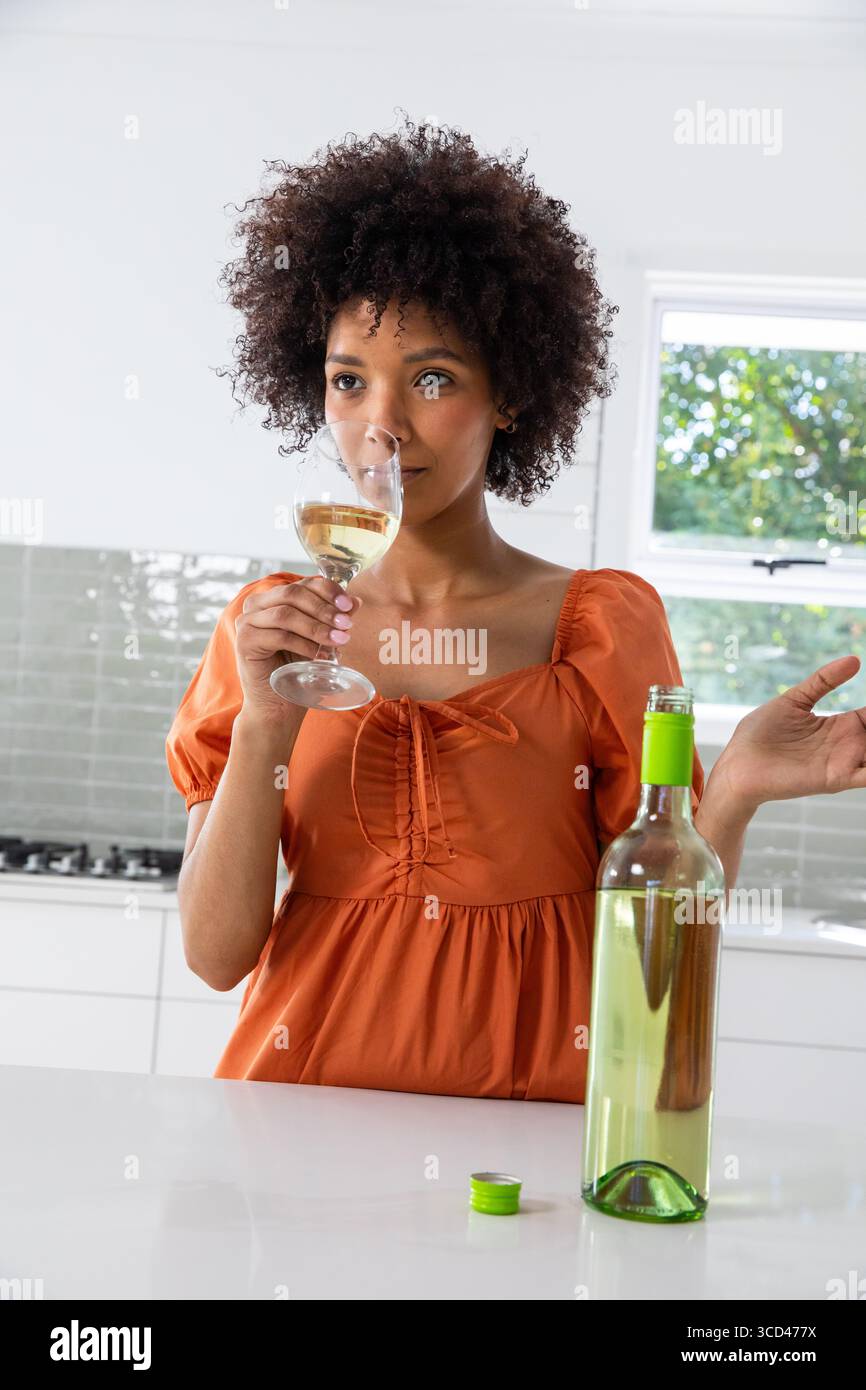 Femme afro-américaine portant une robe orange reniflant un verre de vin blanc à l'îlot de cuisine Banque D'Images