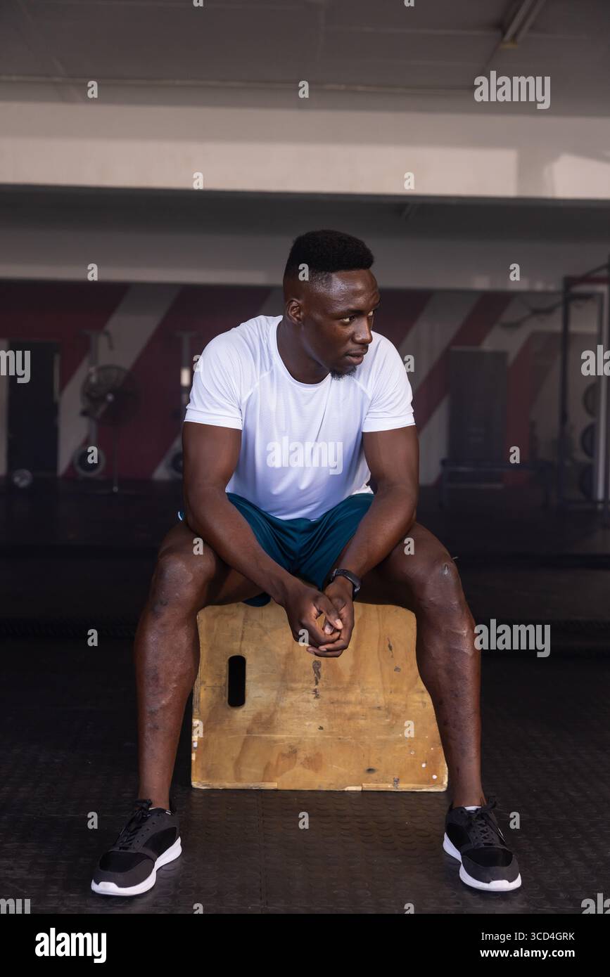 Homme afro-américain reposant sur une boîte plyométrique au gymnase à côté d'appareils de musculation et de tapis en caoutchouc Banque D'Images