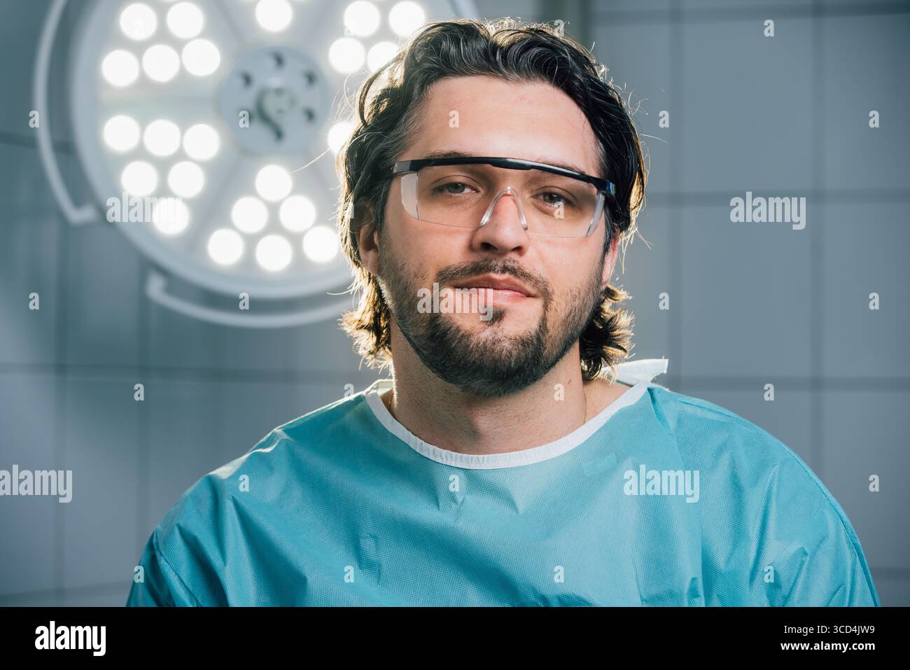 Chirurgien masculin dans les gommages chirurgicaux et les lunettes de protection, se tient en confiance dans une salle d'opération avec des lumières chirurgicales brillantes, soulignant l'engagement à Banque D'Images
