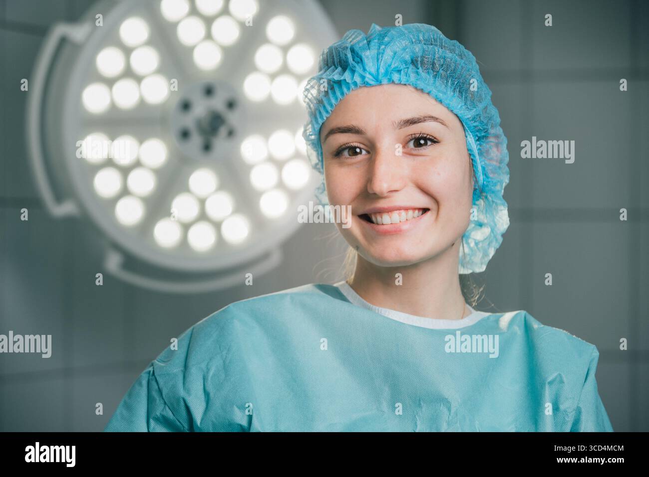Chirurgien féminin en tenue chirurgicale bleue, souriant dans une salle d'opération avec des lumières chirurgicales brillantes, illustrant un engagement envers les soins de santé et le patient Banque D'Images