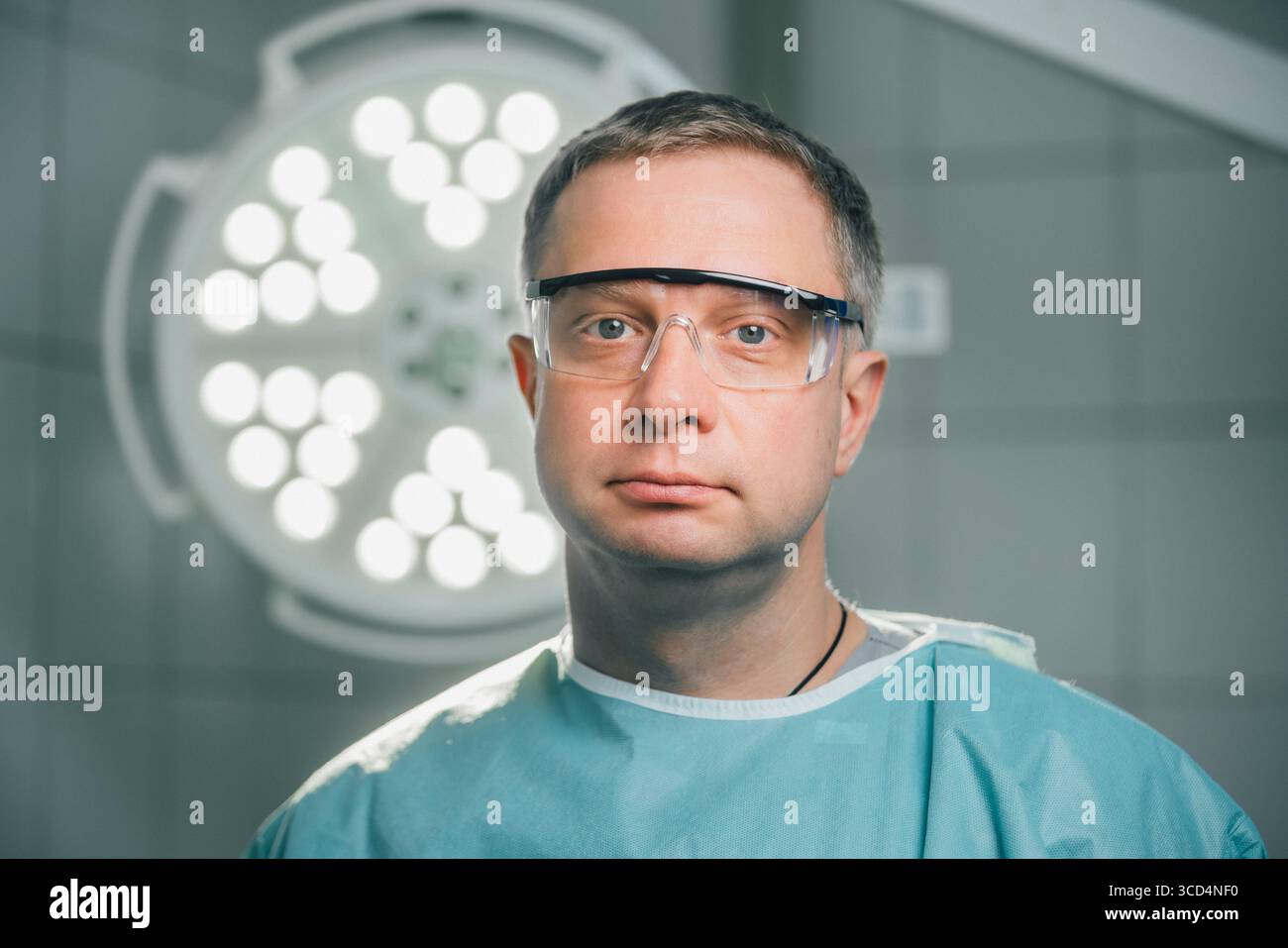 Chirurgien masculin dans les gommages et les lunettes, se tient dans une salle d'opération bien éclairée remplie d'équipement médical de pointe, se préparant à un processus chirurgical critique Banque D'Images