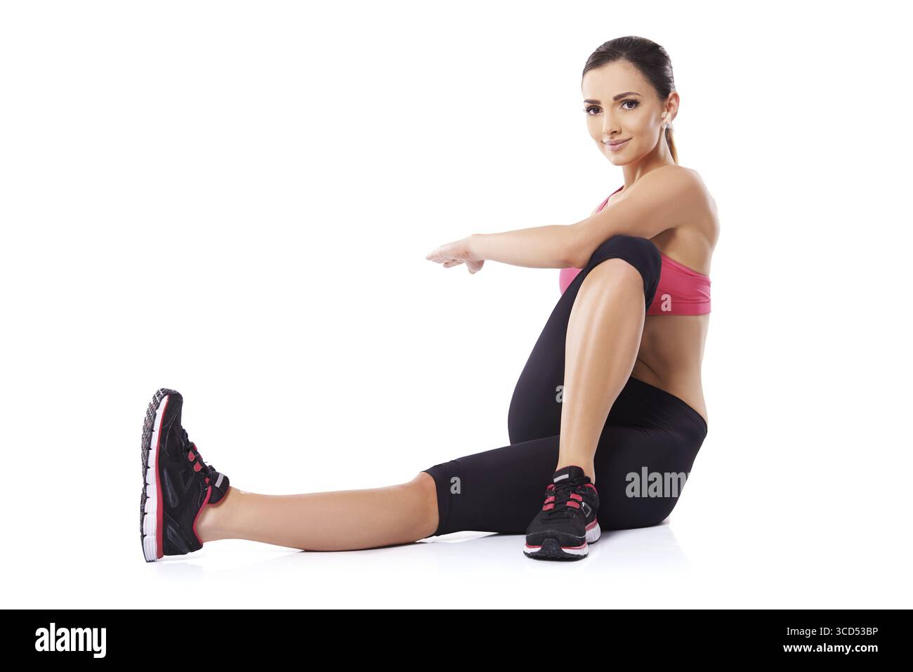 Belle femme souriante assise sur le sol faisant des exercices de torsion du corps en faisant tourner son bras et sa jambe dans des directions opposées, sur blanc Banque D'Images