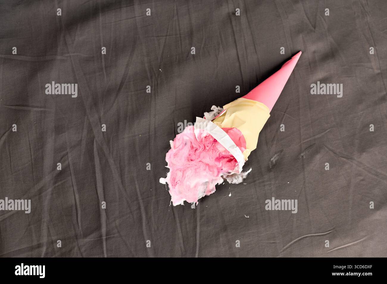 Cône de crème glacée rose couché latéralement sur une surface sombre, partiellement écrasé. L'éclairage projette des ombres douces, mettant en évidence le rose vif contre le gris neutre bac Banque D'Images