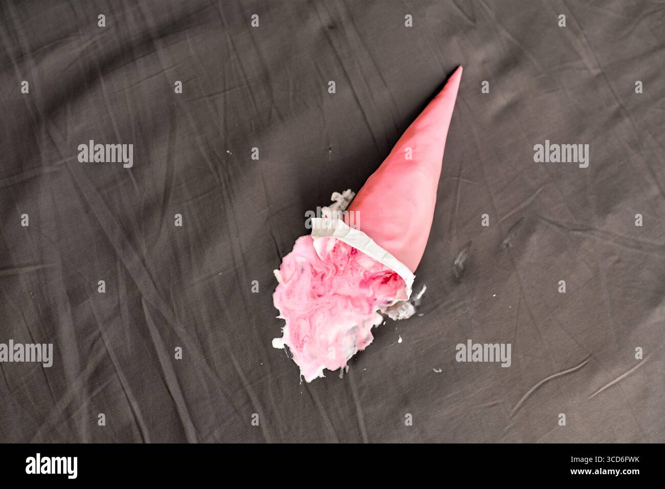 Cône de crème glacée rose couché latéralement sur une surface sombre, partiellement écrasé. L'éclairage projette des ombres douces, mettant en évidence le rose vif contre le gris neutre bac Banque D'Images