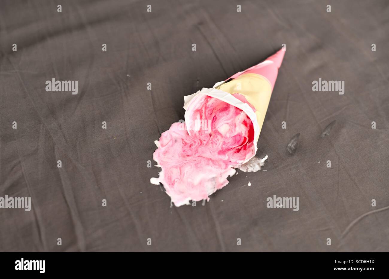 Cône de crème glacée rose couché latéralement sur une surface sombre, partiellement écrasé. L'éclairage projette des ombres douces, mettant en évidence le rose vif contre le gris neutre bac Banque D'Images