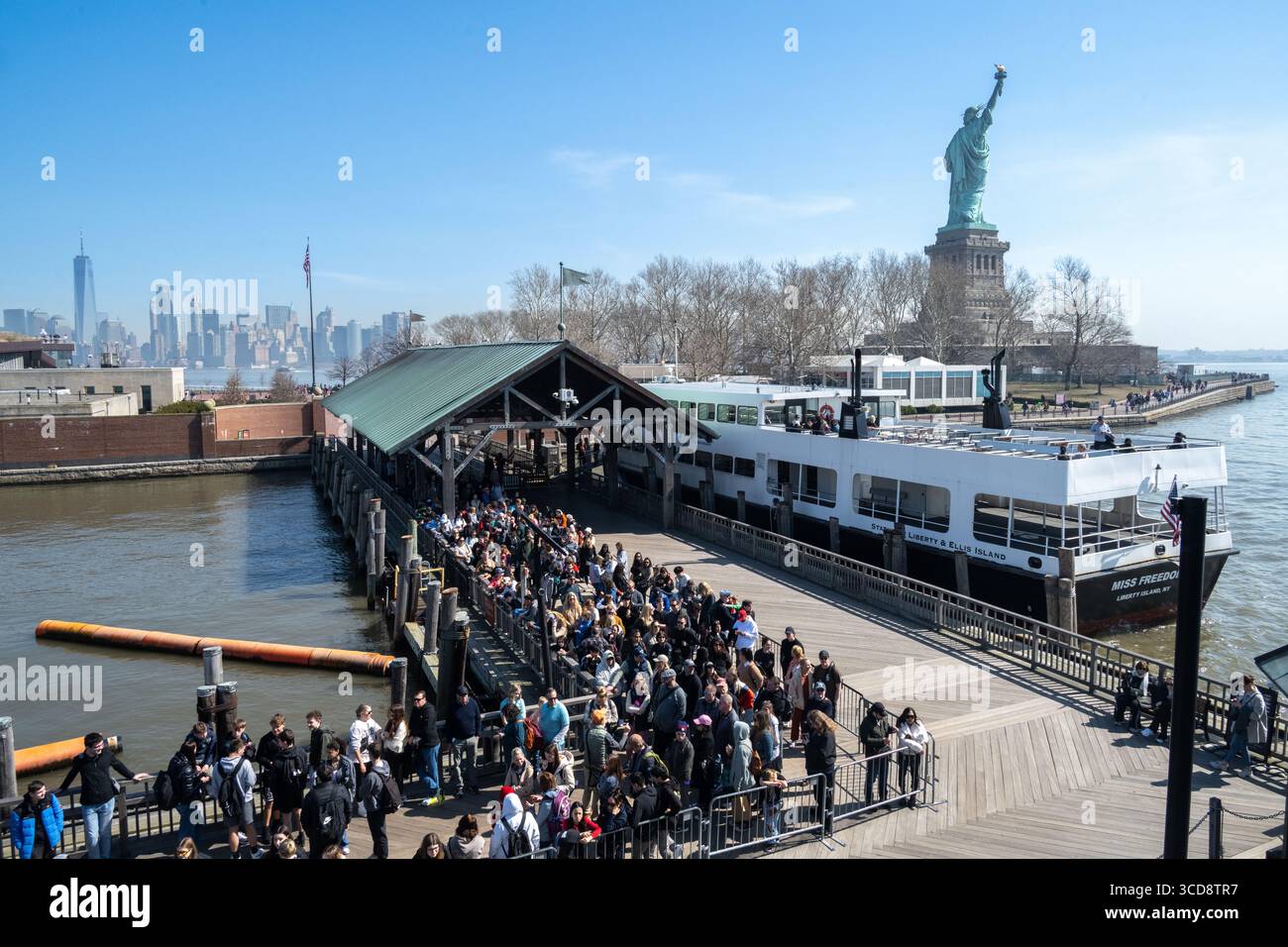 Vue depuis le bateau avant de sortir pour entrer dans Liberty Island avec des visiteurs attendant d'entrer dans le bateau avec Manhattan au loin Banque D'Images
