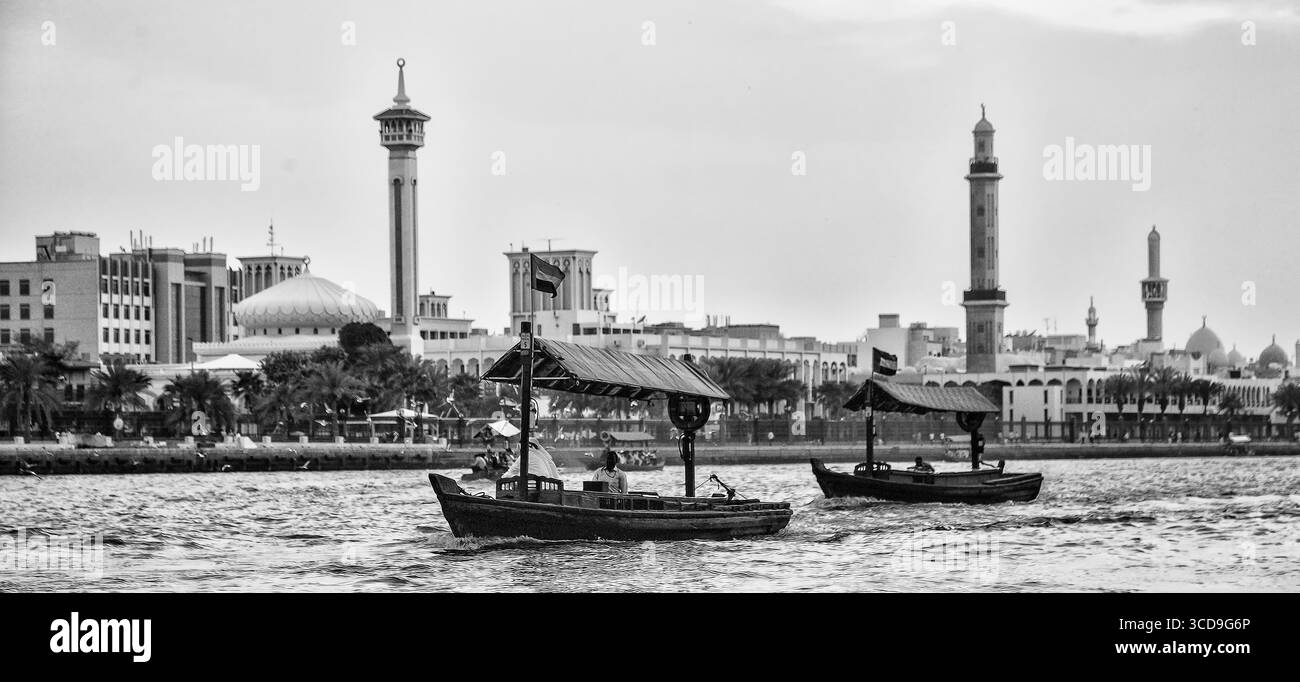 Des bateaux abra traditionnels en bois naviguant sur le Dubai Creek en noir et blanc avec les minarets d'une mosquée historique le long du front de mer. Banque D'Images