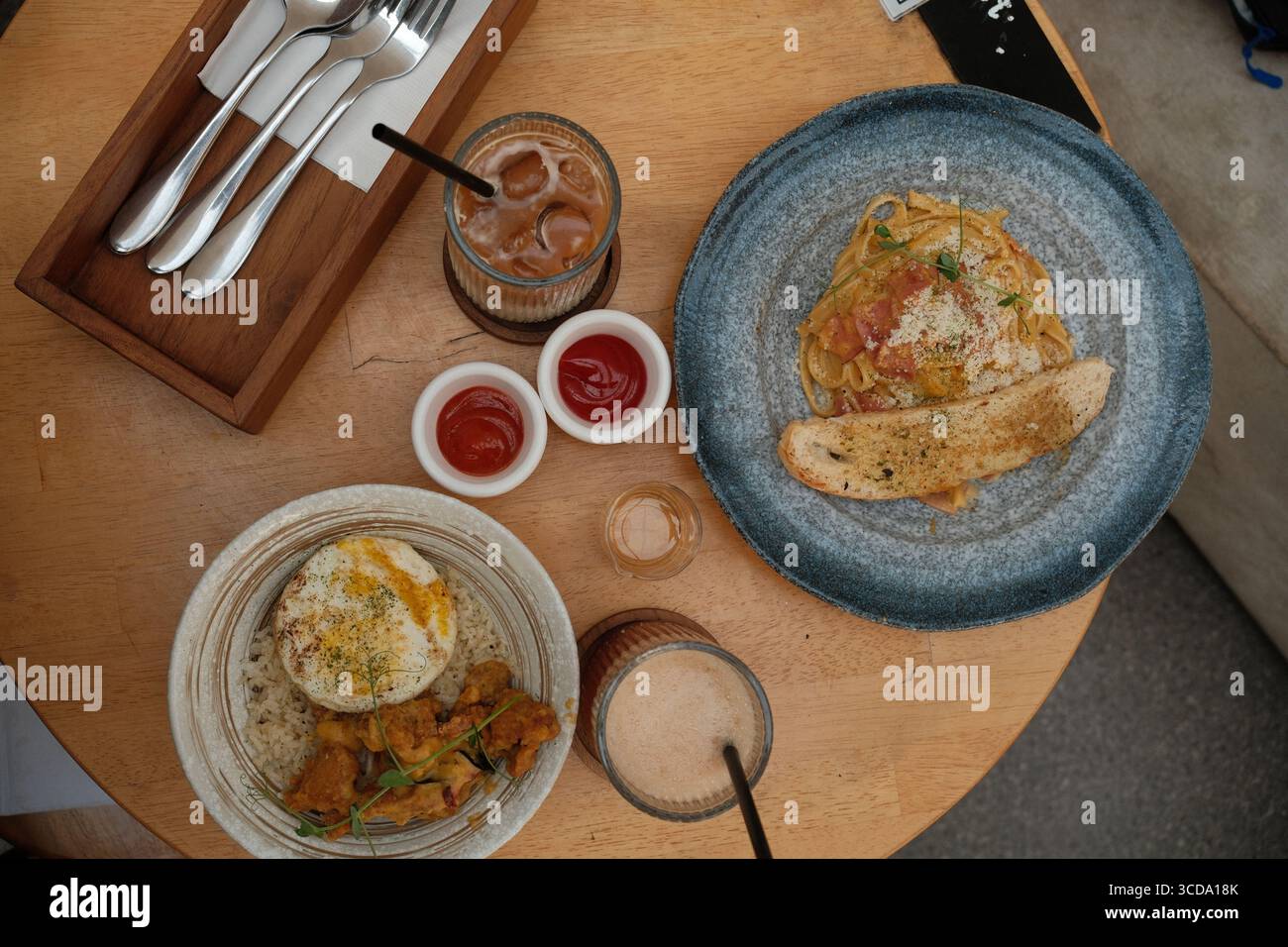 Vue de dessus des pâtes avec pain à l'ail, riz avec poulet frit et oeuf, boissons glacées et condiments servis sur une table de café en bois. Banque D'Images