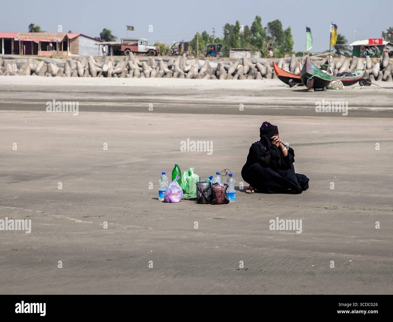 Cox's Bazar, Bangladesh - 08 février 2024 : vue d'une femme en noir, assise sur la vaste plage près de Cox's Bazar - Teknaf Highway, entourée de bouteilles colorées sous un ciel clair. Banque D'Images