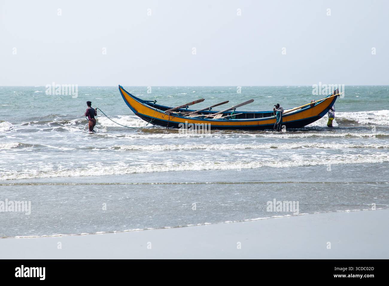 Cox's Bazar, Bangladesh - 08 février 2024 : vue d'un bateau de pêche jaune et bleu éclatant tiré à terre par des pêcheurs locaux, au milieu des vagues à crête blanche. Banque D'Images