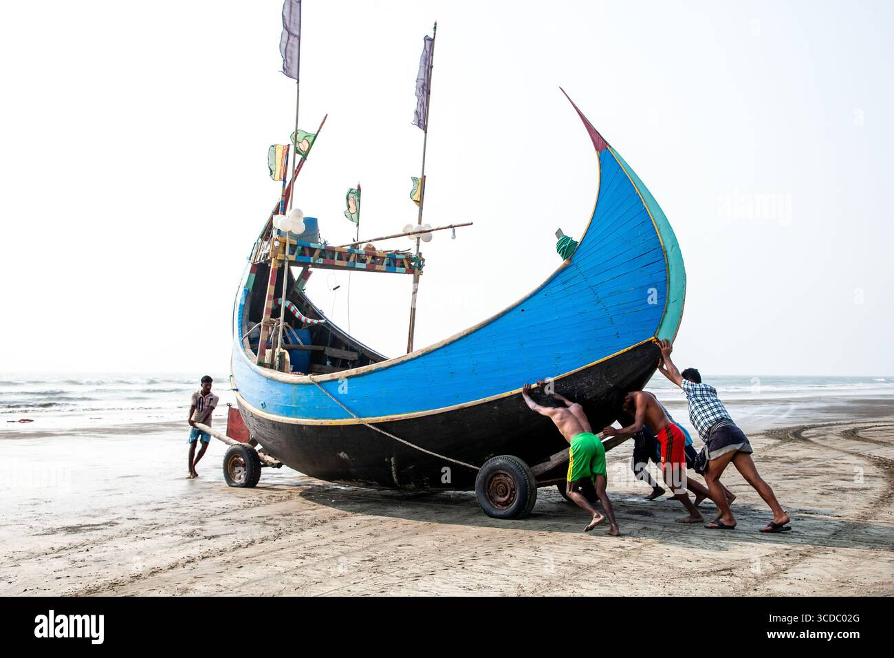 Cox's Bazar, Bangladesh - 06 février 2024 : vue de pêcheurs qui se battent contre le poids, poussant un bateau bleu et noir éclatant à terre sur la route sablonneuse Cox's Bazar - Teknaf. Banque D'Images