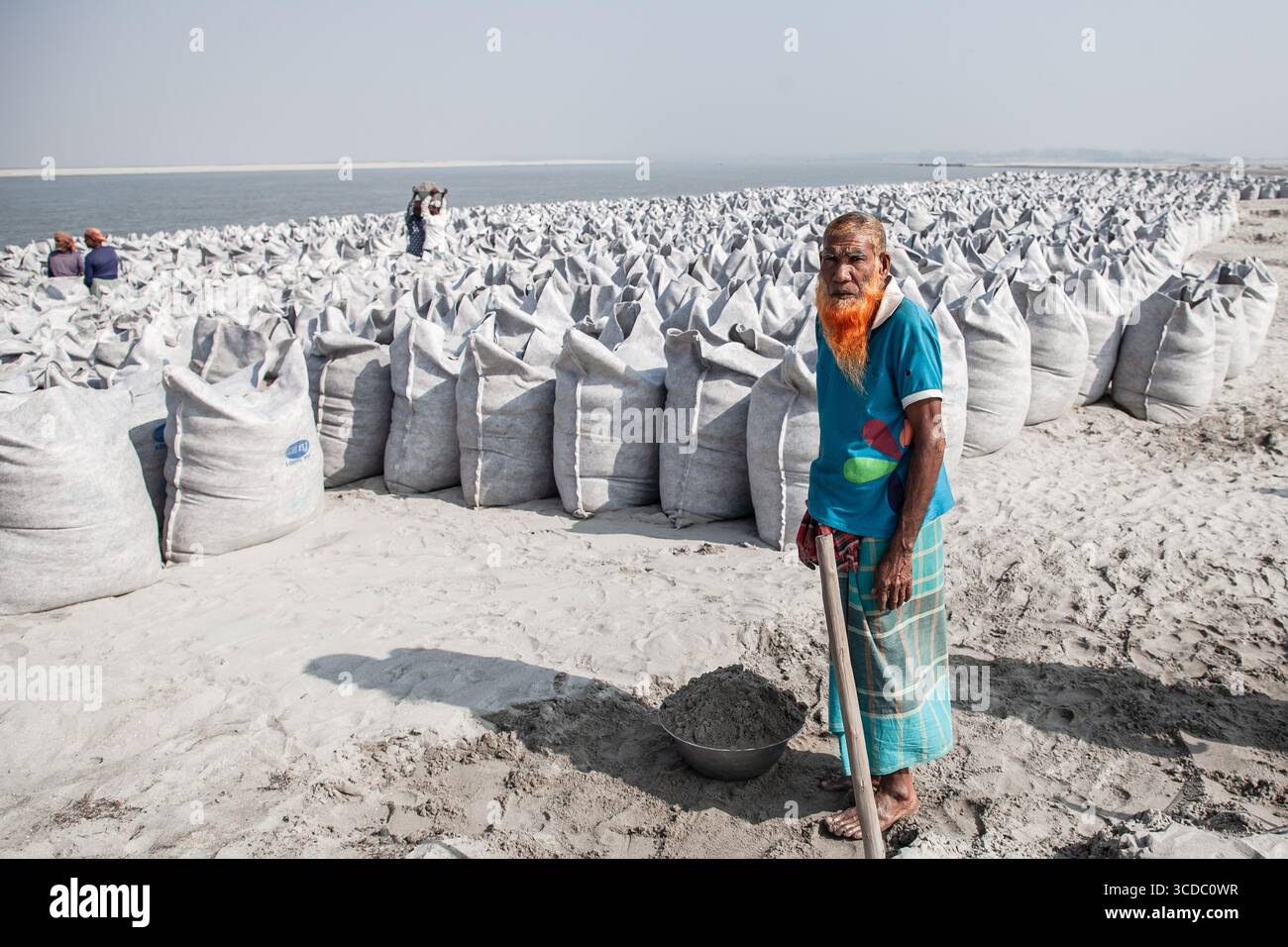 Gaibandha, Bangladesh - 15 décembre 2024 : vue d'un homme à la barbe orange éclatante debout au milieu d'innombrables sacs le long de la rive, sous le ciel vaste et pâle. Banque D'Images