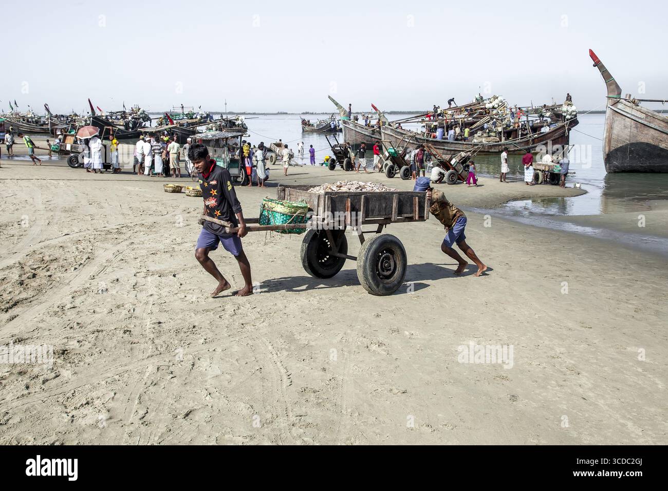 Cox's Bazar, Bangladesh - 29 novembre 2022 : vue d'hommes luttant pour tirer une charrette lourde à travers la plage de sable, avec des bateaux de pêche groupés le long du rivage sous un ciel brumeux. Banque D'Images
