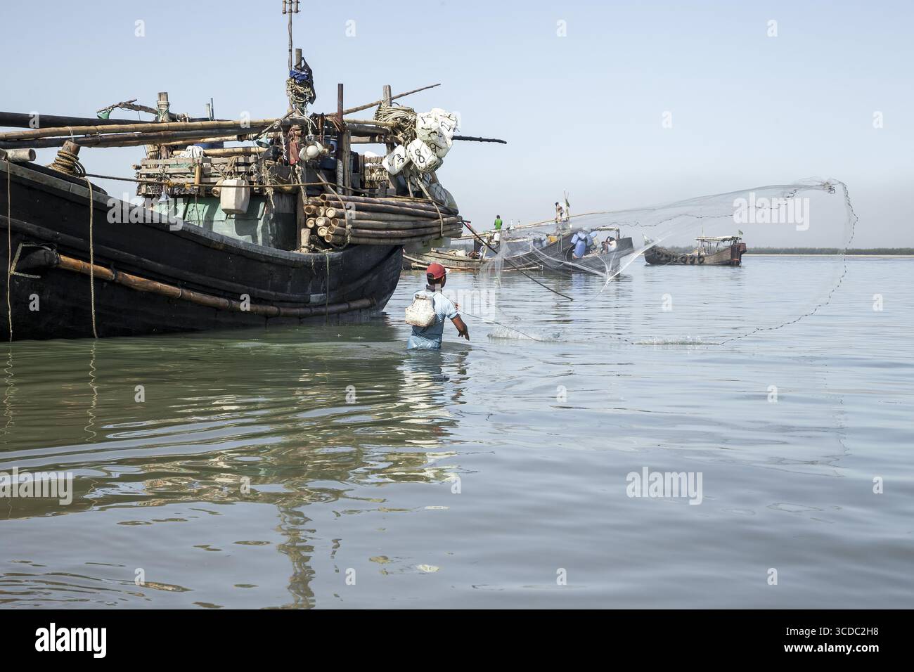 Cox's Bazar, Bangladesh - 29 novembre 2022 : vue d'un pêcheur solitaire jetant son filet dans les eaux chatoyantes, des bateaux traditionnels parsemant l'horizon sous un ciel clair. Banque D'Images
