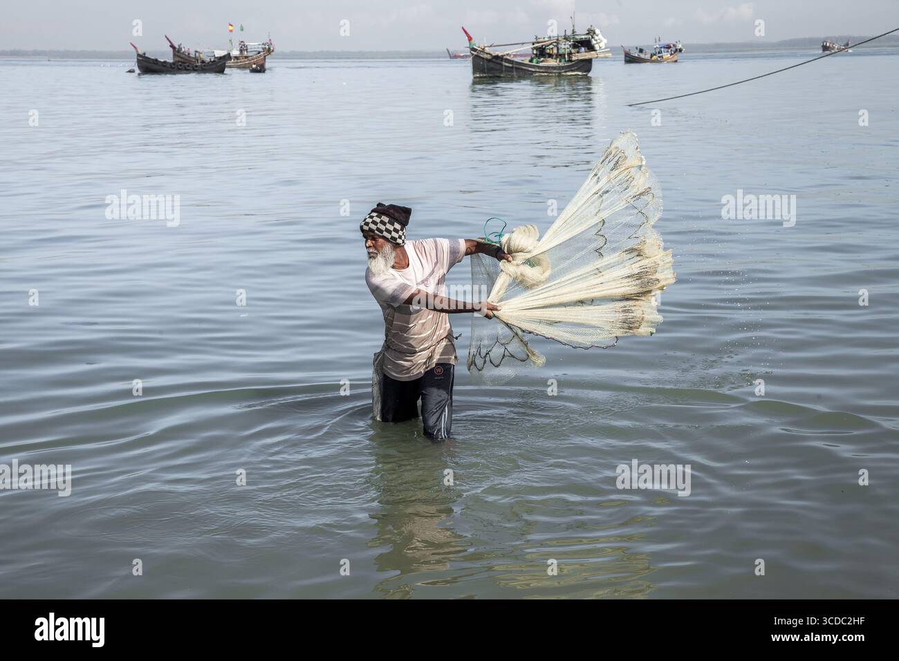 Cox's Bazar, Bangladesh - 29 novembre 2022 : vue d'un pêcheur au fond de la taille dans les eaux sereines et ondulantes, jetant un filet, avec des bateaux de pêche silhouettes contre l'horizon. Banque D'Images