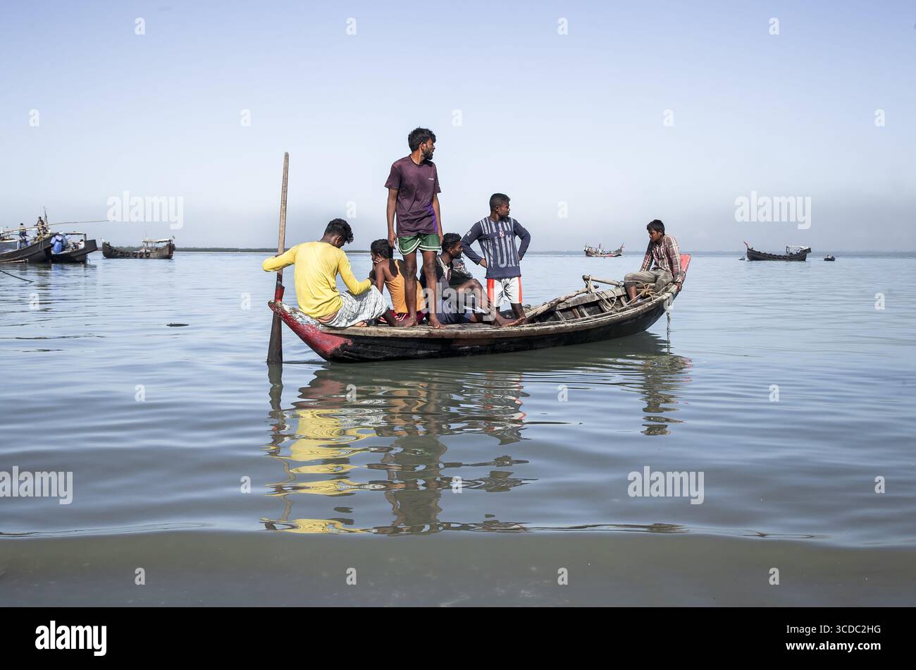 Cox's Bazar, Bangladesh - 29 novembre 2022 : vue de garçons locaux regroupés sur un bateau traditionnel en bois, son reflet chatoyant sur les eaux tranquilles, sous un ciel clair et large. Banque D'Images