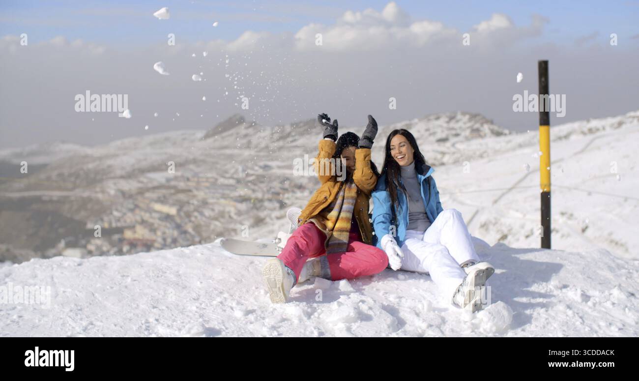 Deux jeunes femmes s'amusant dans la neige hivernale alors qu'elles s'assoient au bord d'une piste de ski dans une station de montagne jetant des boules de neige en l'air et riant Banque D'Images