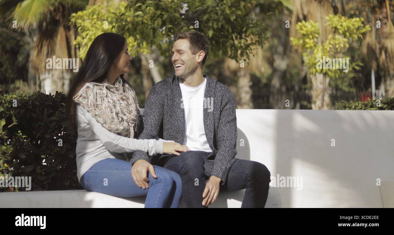 Jeune couple affectueux profitant d'une conversation tranquille ensemble assis sur un mur dans un parc urbain au soleil avec espace de copie Banque D'Images