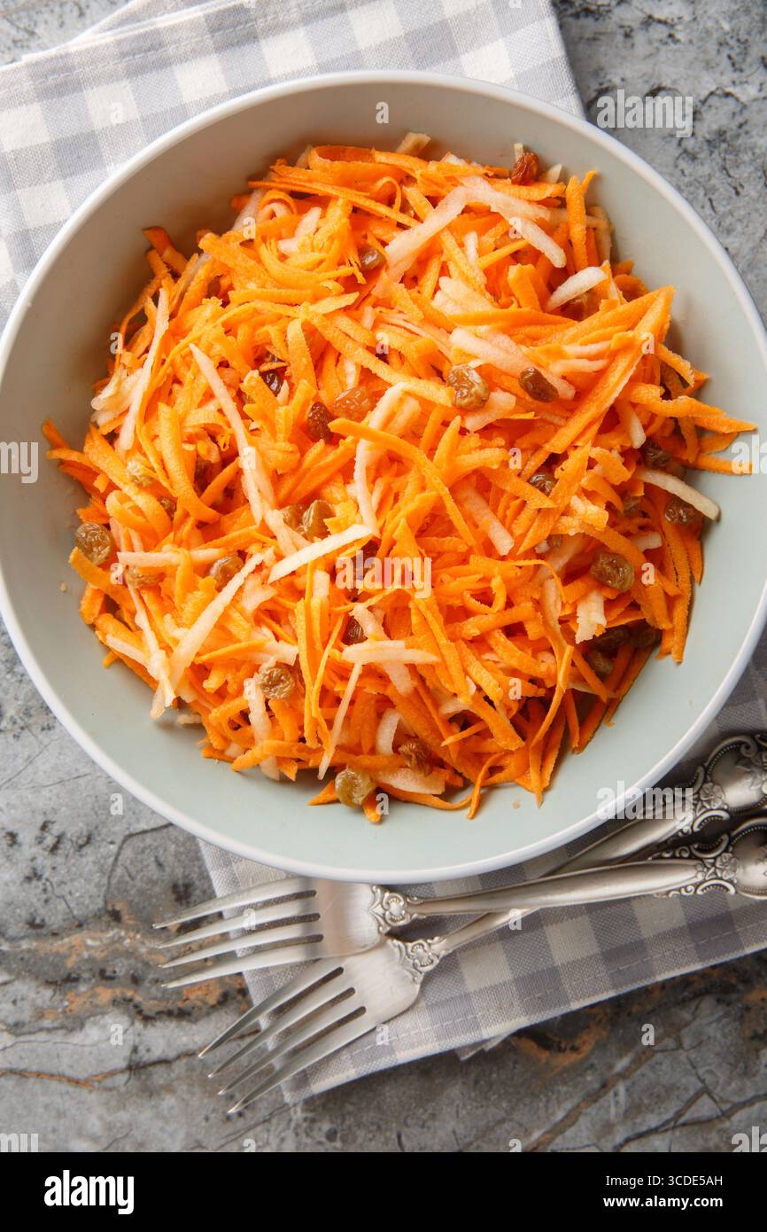 Délicieuse salade de pommes carottes maison avec raisins secs avec vinaigrette au citron gros plan dans un bol sur la table. Vue de dessus verticale Banque D'Images