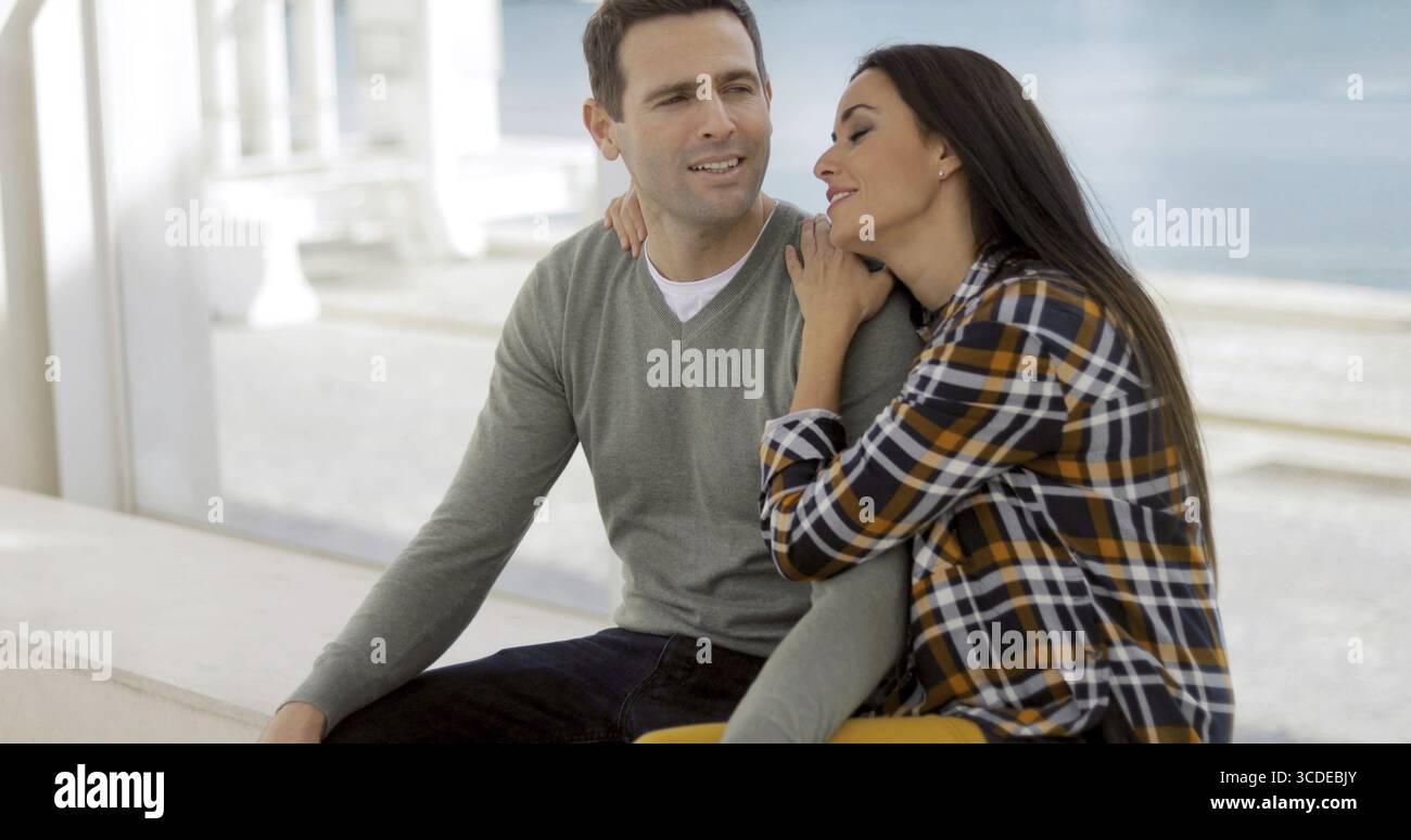 Couple assis dans les bras sur une promenade de bord de regarder quelque chose sur le côté droit du cadre avec un sourire Banque D'Images